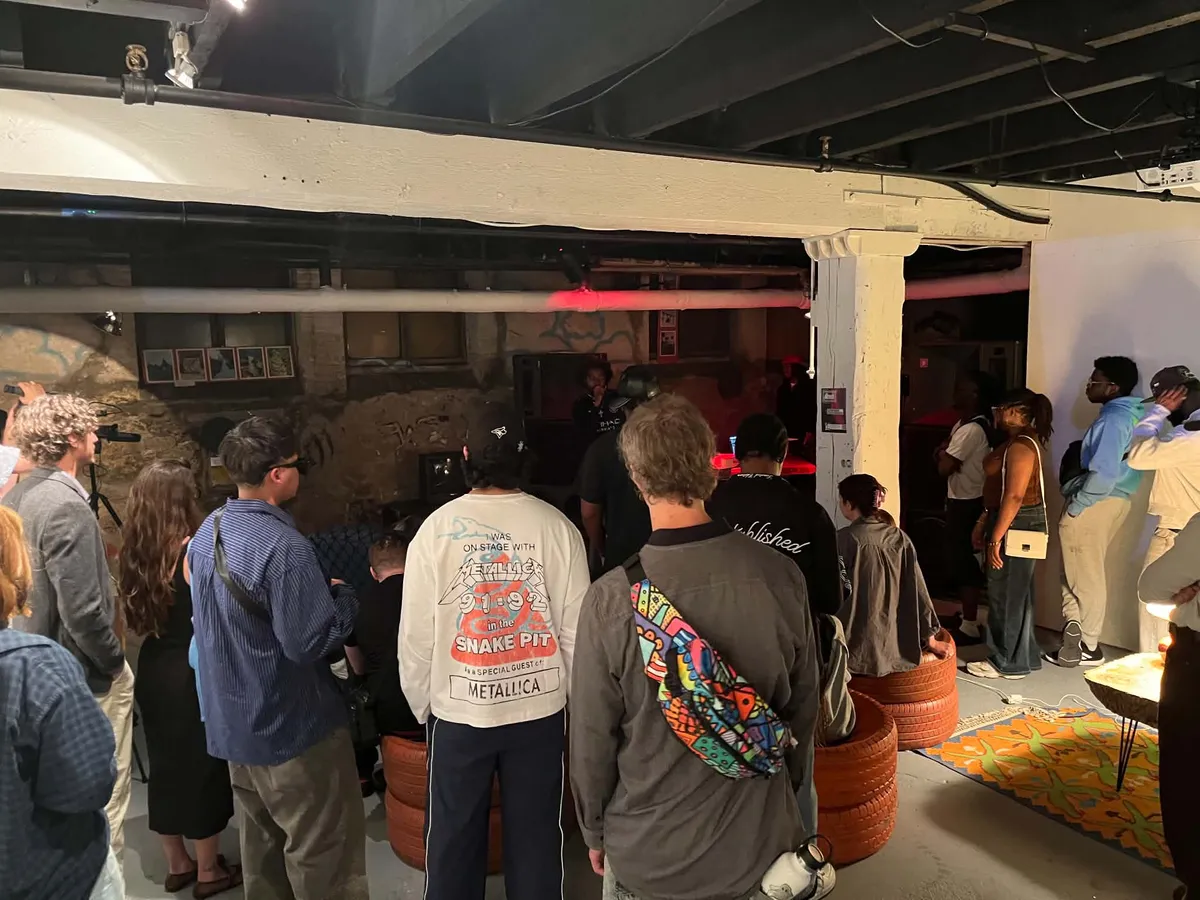An audience standing and sitting on stacked tires, watching a performer at the far end of the basement.