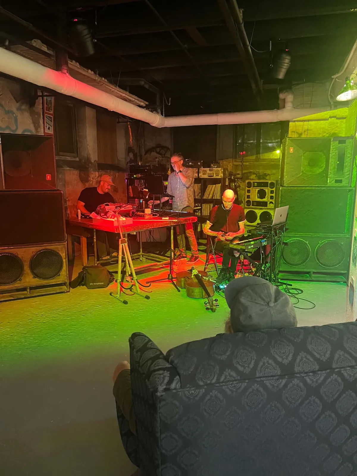 Three musicians playing electronics and keyboards between stacks of speaker cabinets, lit red and green.