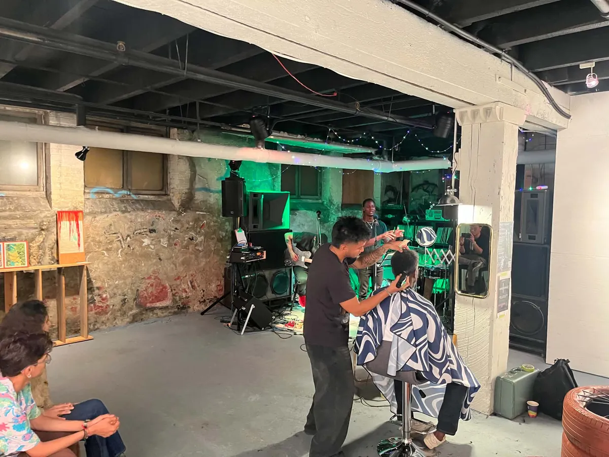 A barber cutting someone’s hair on a stool while a band plays behind them in a green-lit basement, paintings propped against the brick wall.